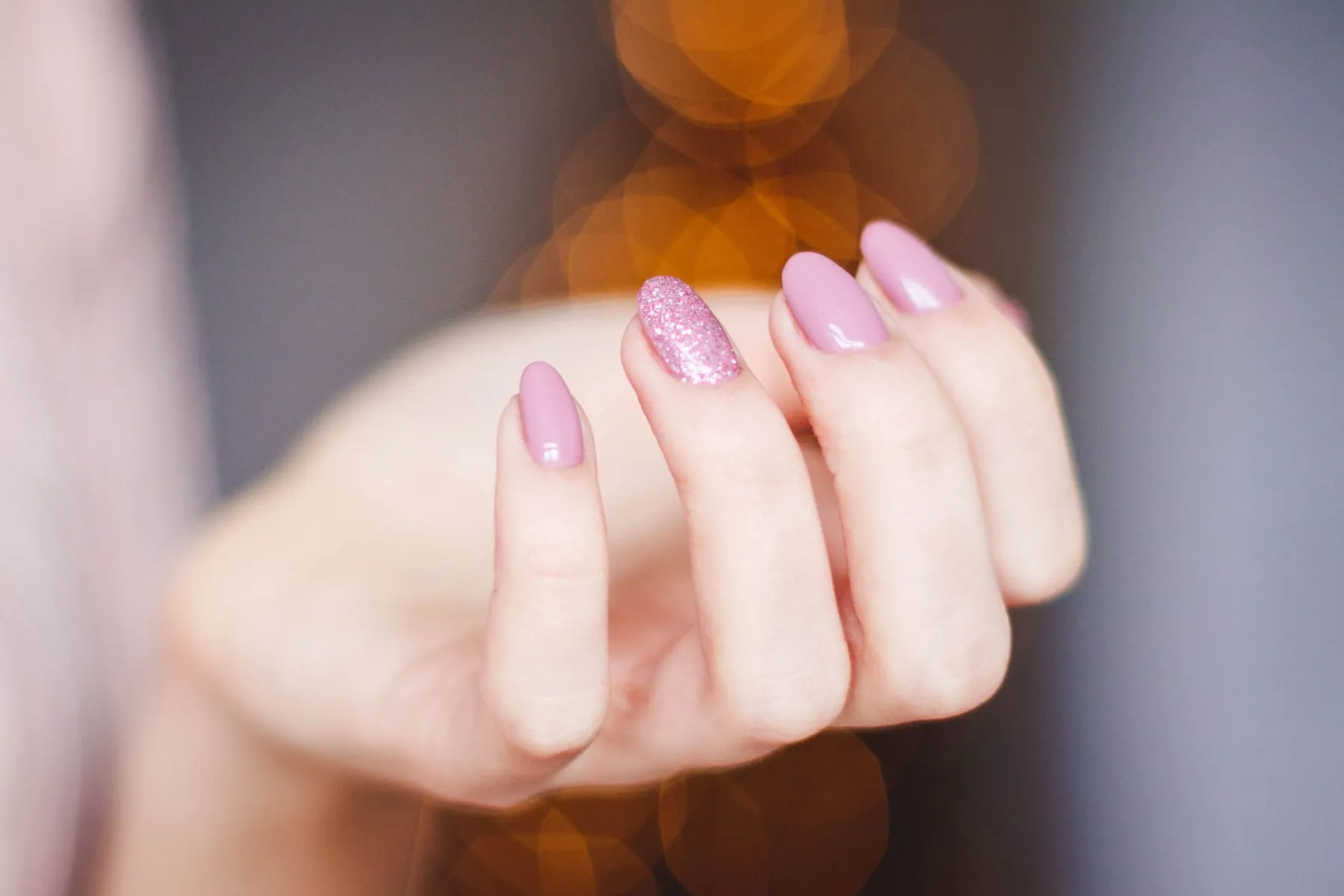 A hand with almond-shaped nails in dusty pink gel polish, one nail in pink glitter, against soft bokeh lights