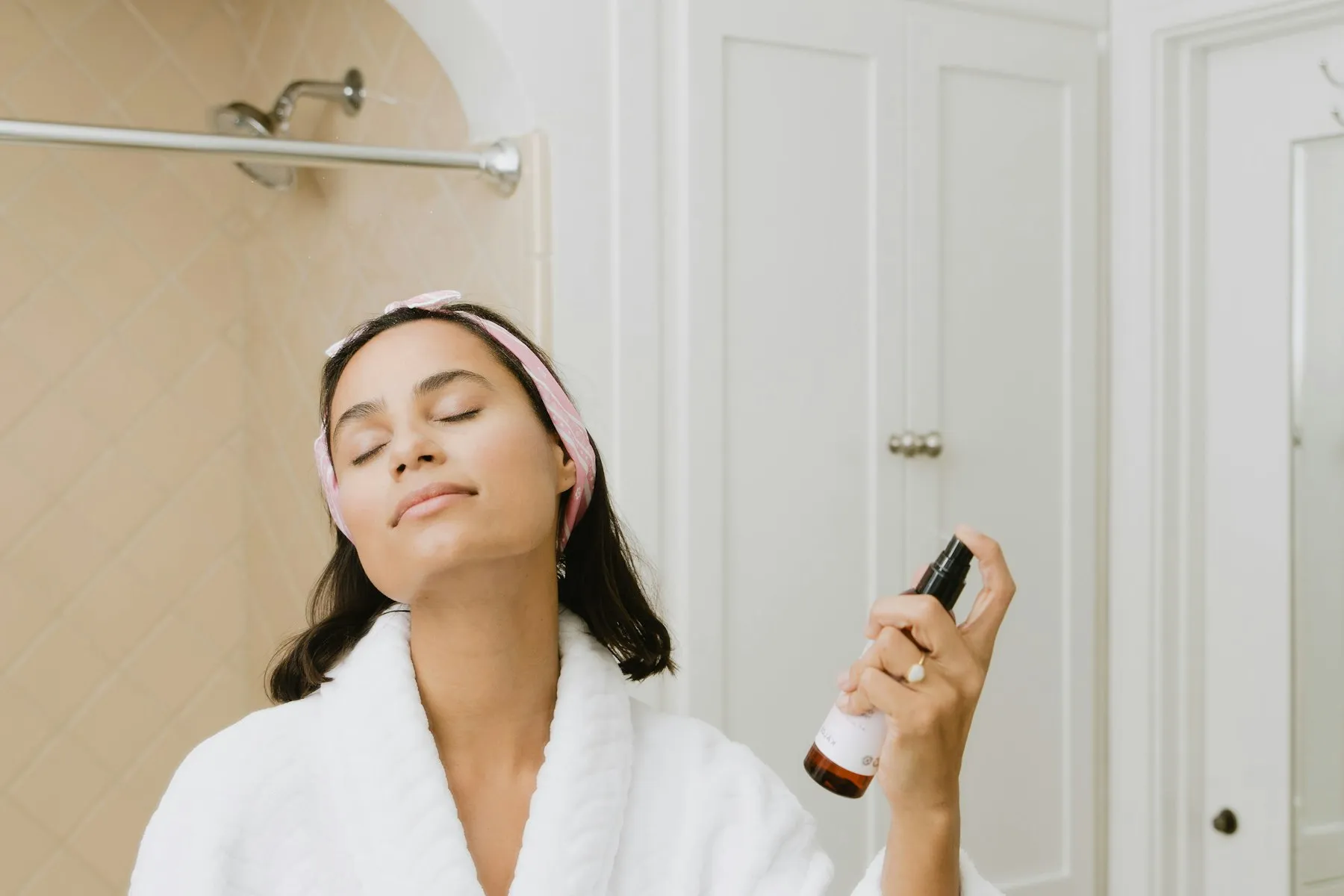 A woman in a white robe spraying facial mist with her eyes closed