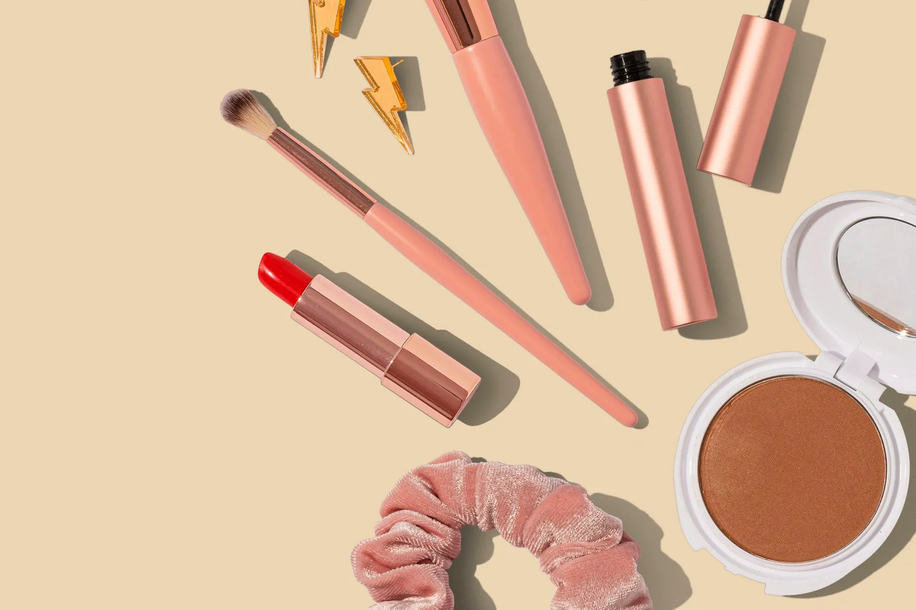 A flat lay of rose-gold makeup brushes, lipsticks and a compact on a warm beige background
